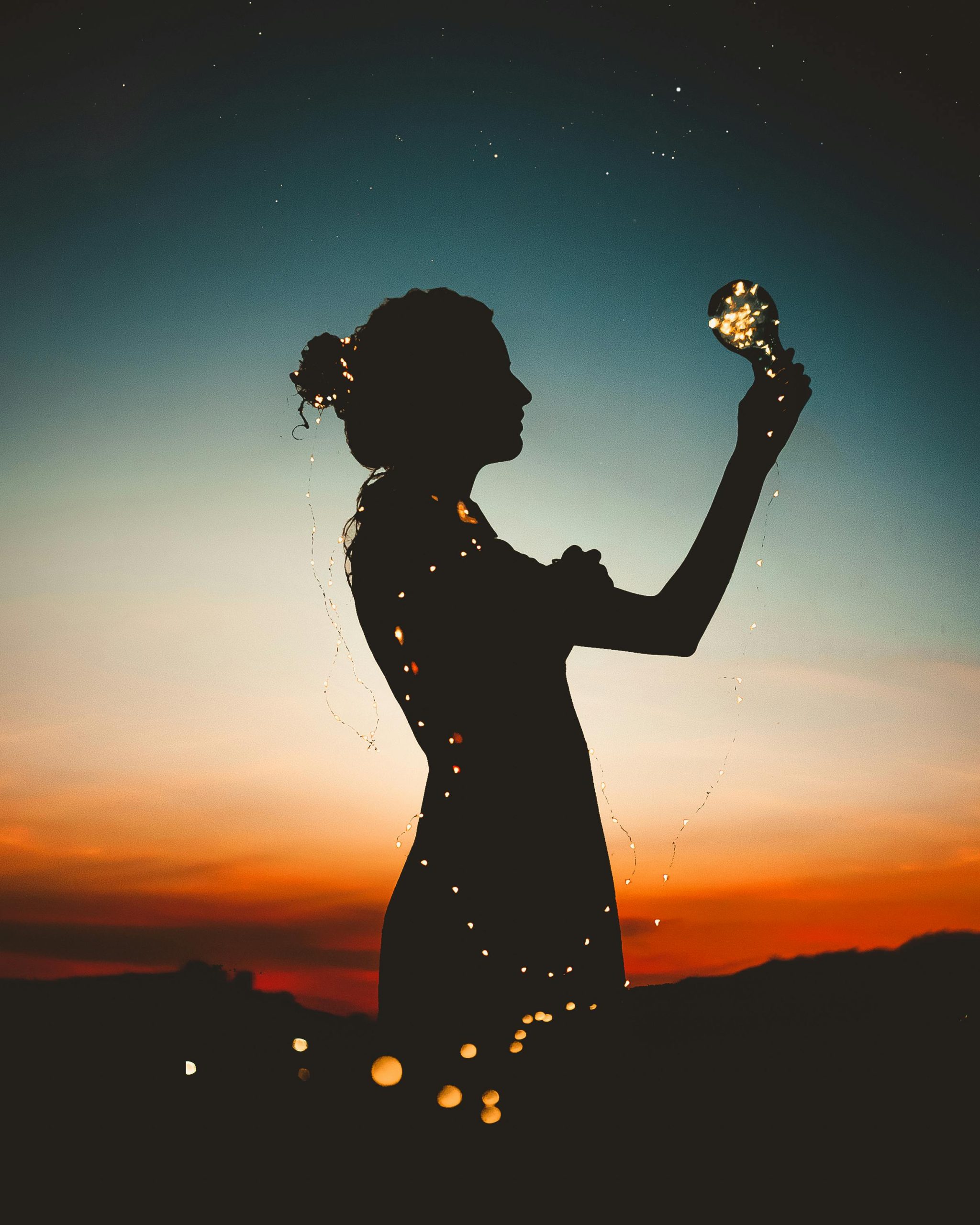 Artistic silhouette of a woman wrapped in fairy lights during a vibrant sunset.