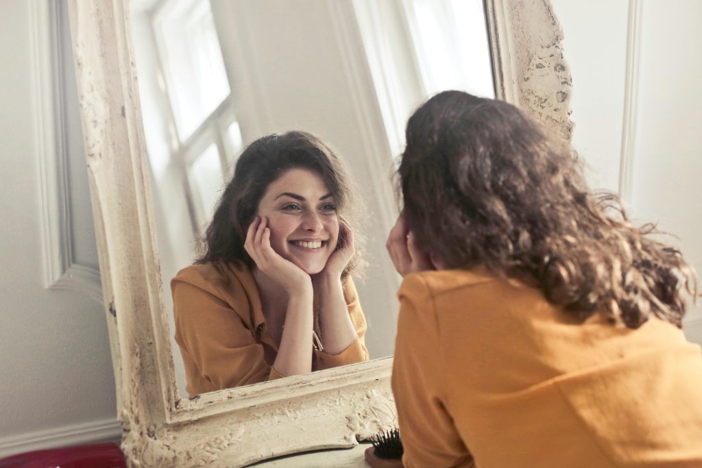 A cheerful woman smiles at her reflection in a vintage-style mirror, exuding positivity and warmth.